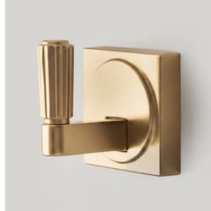 May include: A gold-colored, modern wall hook. The hook features a square backplate with a circular design and a rectangular arm. The hook has a cylindrical, ribbed handle. The hardware is made of metal.