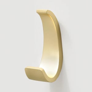 May include: A gold-colored, curved wall hook. The hook has a smooth, matte finish and a simple, modern design. It is mounted on a white wall, providing a minimalist aesthetic. Ideal for hanging coats, bags, or other items.