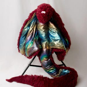 May include: A plush, burgundy-colored creature with a fluffy exterior and iridescent, scale-like interior. The toy has a long, curved tail and a head with small, white eyes. The creature is posed on a black stand against a white backdrop.