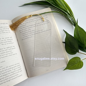 Puede incluir: Un marcapáginas transparente con bordes redondeados y un borla dorada. El marcapáginas está colocado en un libro con texto que dice "Una lluvia de flechas silbó sobre nuestras cabezas mientras caíamos hacia el océano." El marcapáginas tiene el sitio web "snugabee.etsy.com" impreso en él.