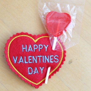 May include: Red felt heart with the text "Happy Valentines Day" embroidered in blue thread. A red heart-shaped lollipop is attached to the heart.