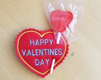 May include: Red felt heart with the text "Happy Valentines Day" embroidered in blue thread. A red heart-shaped lollipop is attached to the heart.