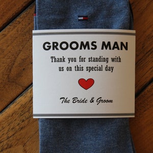 May include: A white card with black text that reads "Grooms Man" and "Thank you for standing with us on this special day."  The card also has a red heart and the text "The Bride & Groom". The card is wrapped around a pair of blue socks.