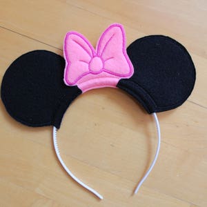 May include: A pink bow headband with black felt ears. The headband is white and the ears are attached to the headband with a white elastic band.