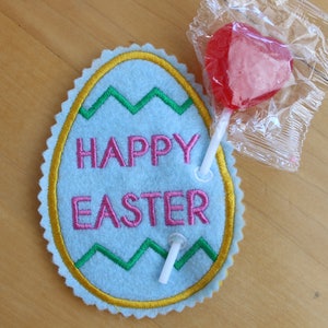 May include: A blue felt Easter egg with a yellow border and green zig-zag design. The egg has the words "Happy Easter" embroidered in pink. Two white lollipop sticks are attached to the egg.