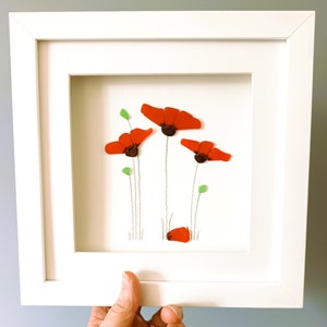 May include: A framed artwork featuring a cluster of red poppies made from sea glass. The poppies are arranged in a simple, elegant design with green stems and leaves.