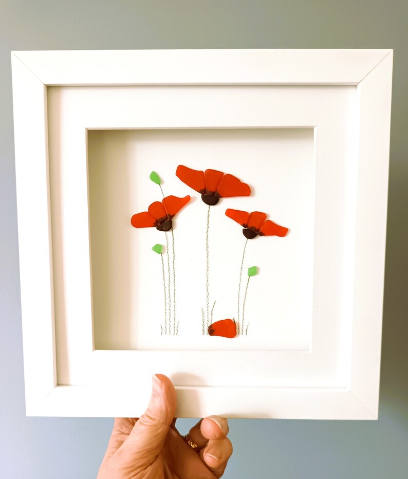 May include: A framed artwork featuring a cluster of red poppies made from sea glass. The poppies are arranged in a simple, elegant design with green stems and leaves.