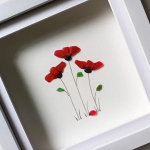 May include: Framed artwork featuring three red poppies made from sea glass, with green leaves and stems drawn in pencil on a white background.