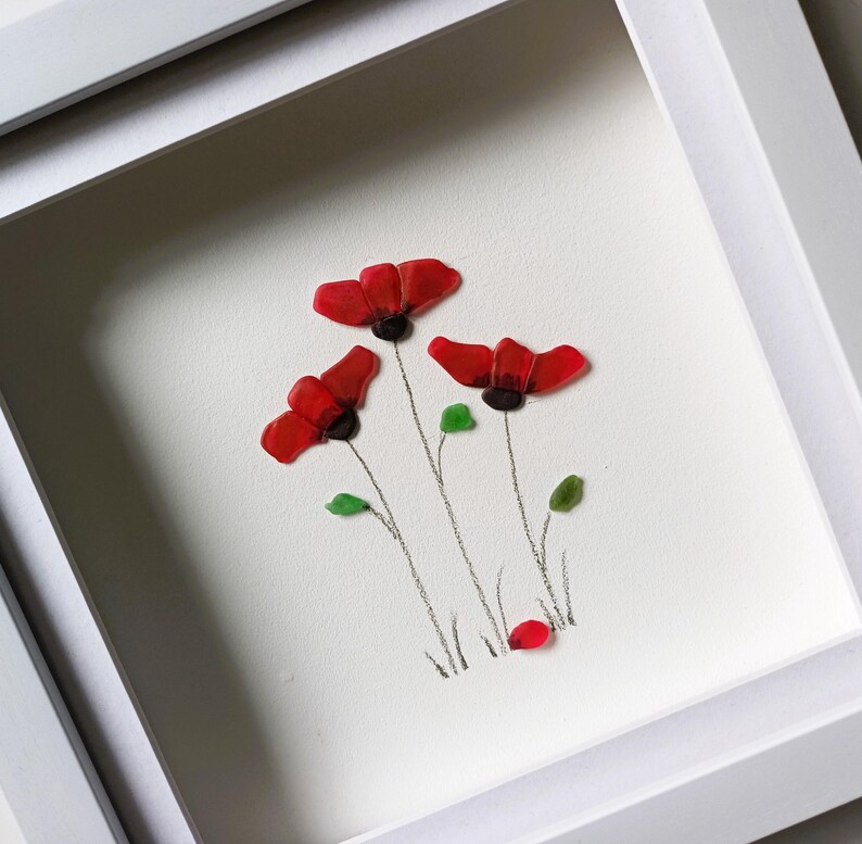 May include: Framed artwork featuring three red poppies made from sea glass, with green leaves and stems drawn in pencil on a white background.