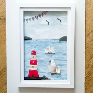 May include: Framed artwork showing a coastal scene. A red and white lighthouse is in the foreground, with sailboats on the blue water. The sky is light blue with seagulls and a banner of flags.