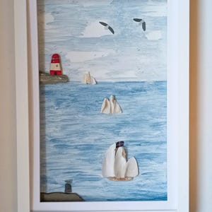 May include: Framed artwork of a coastal scene with a red and white lighthouse, sailboats, and seagulls. The painting uses blue and white tones to depict the sea and sky. The artwork is presented in a white frame.
