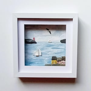 May include: A framed artwork showing a coastal scene. The image features a blue sea with sailboats, a lighthouse, and small houses. A seagull flies in the sky. The frame is white.