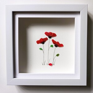 May include: A framed artwork featuring a cluster of red poppies made from sea glass. The poppies are arranged in a simple, elegant design with green leaves and stems. The artwork is set against a white background and is framed in a white frame.
