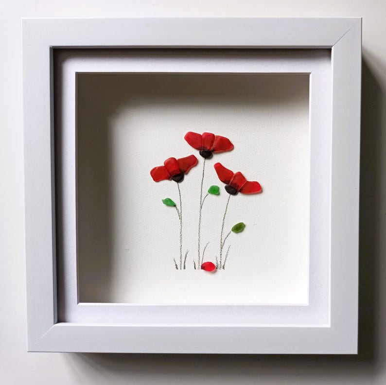 May include: A framed artwork featuring a cluster of red poppies made from sea glass. The poppies are arranged in a simple, elegant design with green leaves and stems. The artwork is set against a white background and is framed in a white frame.