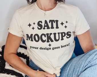 May include: A woman wearing a cream-colored t-shirt with the text "SATI MOCKUPS your design goes here!" in black. She is wearing blue jeans and has her hand on the front of the shirt.