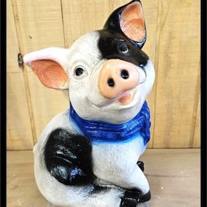 May include: A ceramic pig figurine with a white body, black patches, and a blue scarf. The pig has pink ears and snout, and is sitting upright. The figurine is a decorative item.