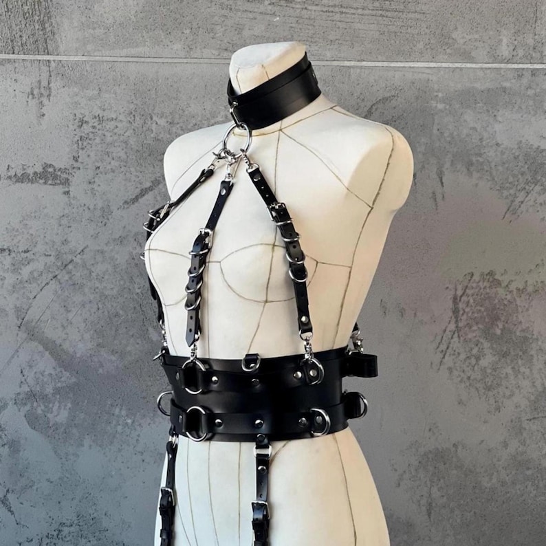 May include: Black leather body harness with a choker collar, straps, and a wide belt. The harness features silver-toned metal rings and buckles. The design is displayed on a mannequin against a gray background.