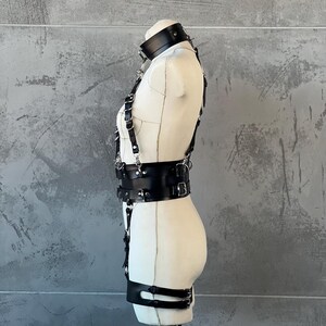 May include: Black leather harness with silver-tone hardware. The harness includes a neck collar, chest straps, and a waist belt. The design features multiple buckles and adjustable straps. The harness is displayed on a mannequin.