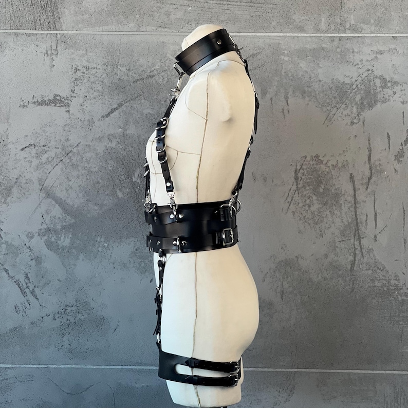May include: Black leather harness with silver-tone hardware. The harness includes a neck collar, chest straps, and a waist belt. The design features multiple buckles and adjustable straps. The harness is displayed on a mannequin.