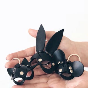 Può includere: Tre portachiavi in pelle nera con design ispirati agli animali. Un portachiavi ha orecchie lunghe e appuntite, un altro ha orecchie arrotondate e il terzo ha orecchie da gatto. Ogni pezzo è realizzato con rivetti e anelli in metallo argentato.