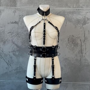 May include: Black leather body harness with a choker collar, chest straps, and a waist belt. The harness features multiple metal rings and adjustable straps. The design includes leg straps with metal hardware, displayed on a mannequin against a gray background.