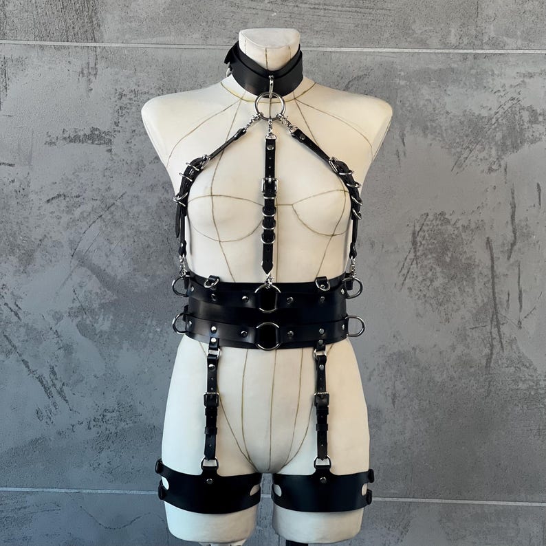 May include: Black leather body harness with a choker collar, chest straps, and a waist belt. The harness features multiple metal rings and adjustable straps. The design includes leg straps with metal hardware, displayed on a mannequin against a gray background.
