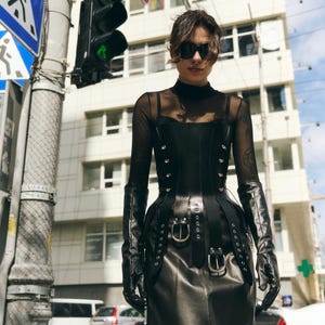 Può includere: Una persona indossa un corsetto in pelle nera con rivetti e fibbie argentate, abbinato a lunghi guanti neri e una gonna nera. L'outfit è ambientato su uno sfondo urbano con un edificio e semafori.