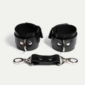May include: Black leather wrist restraints with silver-tone metal hardware. The set includes two cuffs and a connecting strap with two clasps. The cuffs have a buckle closure and are secured with silver rivets.