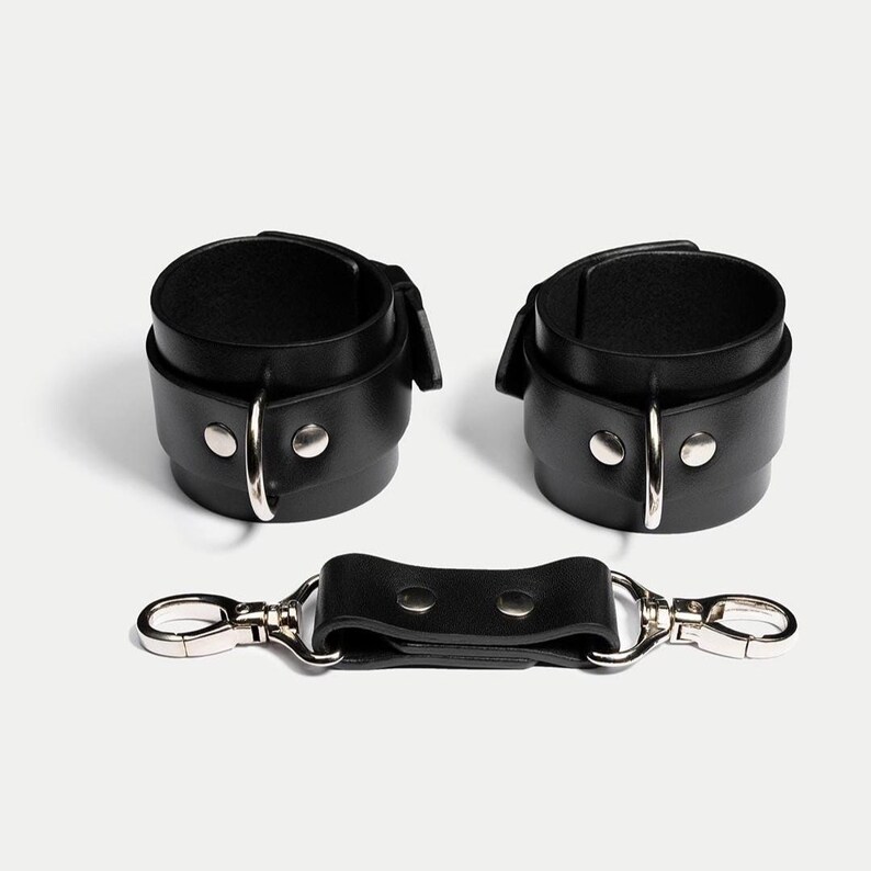 May include: Black leather wrist restraints with silver-tone metal hardware. The set includes two cuffs and a connecting strap with two clasps. The cuffs have a buckle closure and are secured with silver rivets.