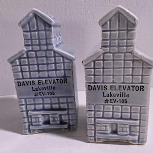 May include: Two gray ceramic salt and pepper shakers shaped like buildings. Each shaker has a rectangular body with a gabled roof and the text "DAVIS ELEVATOR Lakeville #EV-105". The shakers have a grid-like pattern.