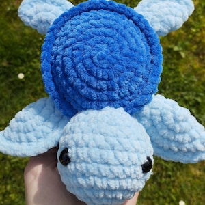 Pode incluir: Um brinquedo de pelúcia de tartaruga marinha feito em crochê, com uma carapaça circular azul vibrante com um padrão espiral texturizado. A cabeça e as quatro nadadeiras são feitas de fio azul claro e tem dois olhos pretos pequenos.