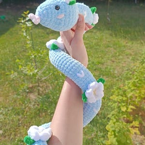 Peut inclure: Un long serpent en peluche crocheté bleu clair avec une finition en fil doux et texturé. Le serpent est décoré de plusieurs fleurs crochetées blanches avec des feuilles vertes attachées le long du corps et de la tête, et possède de petits yeux noirs.