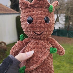 Puede incluir: Una muñeca de peluche marrón que se asemeja a un personaje con una cara sonriente, ojos negros grandes y acentos verdes. La muñeca tiene una apariencia texturizada y borrosa y se sostiene contra un fondo borroso al aire libre.