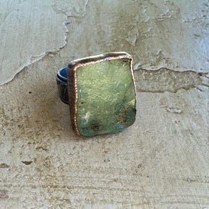 Könnte beinhalten: Ein auffälliger Ring mit einem großen, hellgrünen Stein mit strukturierter Oberfläche. Der Stein ist in einen kupferfarbenen Metallrahmen gefasst. Der Ring hat ein dunkles, gemustertes Band. Der Ring wird auf einer strukturierten, neutralfarbenen Oberfläche gezeigt.