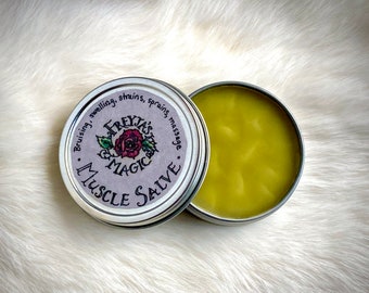 May include: A silver tin with a purple label that reads "Freyja's Magic Muscle Salve" and "Bruising, swelling, strains, sprains, massage". The tin is open and the salve inside is a light yellow color.