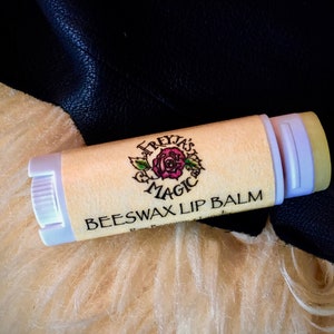 Puede incluir: Un tubo blanco de bálsamo labial de cera de abejas con un diseño de rosa rosa y hojas verdes. La etiqueta dice "Freyja's Magic Beeswax Lip Balm".
