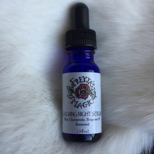 May include: A blue glass bottle with a black dropper top. The bottle is labelled "Freyja's Magic Calming Night Serum Blue Chamomile, Bergamot & Rosewood (.5 fl oz)"