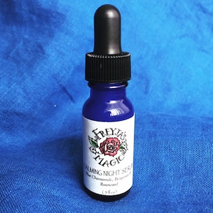 May include: A small blue glass bottle with a black dropper top. The bottle has a white label with the text "Freyja's Magic" and "Calming Night Serum" in black lettering. The label also lists the ingredients: "Blue Chamomile, Bergamot & Rosewood (5 fl oz)"