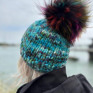Pode incluir: Um gorro de malha feito à mão em tons de azul-turquesa, azul e roxo com um grande pompom de pele sintética multicolorido. O chapéu é feito de fio grosso e é usado por uma pessoa com um casaco preto.