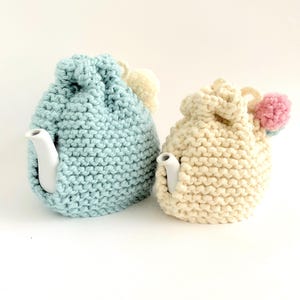 Pode incluir: Dois aquecedores de chaleira de tricô, um em azul claro e outro em creme, com pompons no topo. Os aquecedores são feitos com um ponto de tricô simples e são projetados para manter as chaleiras quentes.