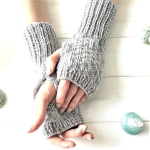 Könnte beinhalten: Ein Paar graue Fingerlose Handschuhe aus gestrickter Wolle. Die Handschuhe sind über den Händen gefaltet und werden vor einem weißen Holz Hintergrund gezeigt.