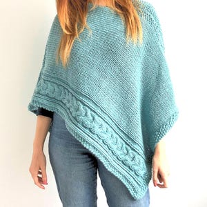 May include: A teal knitted poncho featuring a prominent cable knit border along the bottom edge. The garment has a smooth stockinette stitch texture on the main body and a textured seed stitch finish along the hem and side edges.