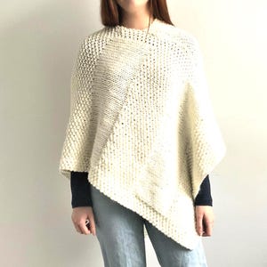 Puede incluir: Un poncho de punto hecho a mano en color blanco roto, que presenta una combinación de paneles con textura en punto bobo y secciones en punto liso. La prenda tiene un cuello redondo y un dobladillo asimétrico que cae en diagonal.