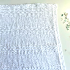 May include: A white knitted baby blanket with a simple stitch pattern. The blanket is folded in half and has a border around the edges.