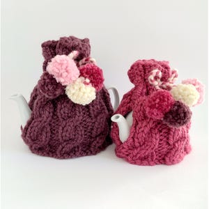 May include: Two knitted teapot cozies in shades of pink and burgundy with fluffy pom-poms. The cozies are made with a cable knit pattern.