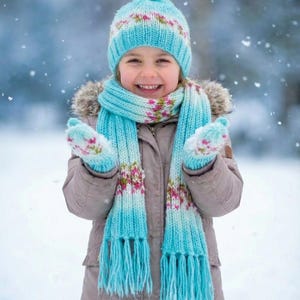 May include: A light blue knit winter set consisting of a pom-pom beanie, a long fringed scarf, and a pair of mittens. Each piece features a ribbed texture and a repeating pattern of small pink and green floral accents throughout the knit fabric.