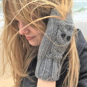 Könnte beinhalten: Graue gestrickte Fingerlose Handschuhe mit einem Zopfmuster und zwei schwarzen Knöpfen am Bündchen.