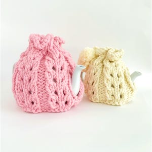 以下が含まれることがあります： ピンクとクリーム色の2つのニットのティーポットカバー。ピンクのカバーから白いティーポットの注ぎ口が出ています。