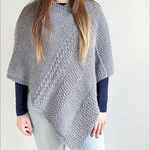 Könnte beinhalten: Ein grauer Strickponcho mit einem diagonalen Muster aus abwechselnden Rippen- und strukturierten Perlmuster-Abschnitten. Das Kleidungsstück hat einen asymmetrischen Saum und einen hohen Ausschnitt, gefertigt aus dickem Garn in einheitlichem Grau.