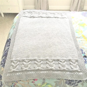 May include: A gray knitted baby blanket with a cable knit border. The blanket is folded and lying on a floral patterned bedspread.